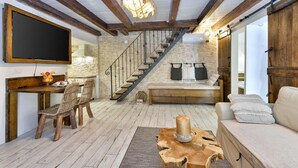 Een rustiek interieur met zichtbare houten balken, een stenen muur, een houten trap, een eethoek met rieten stoelen en een gezellige zithoek met een bank en kussens.