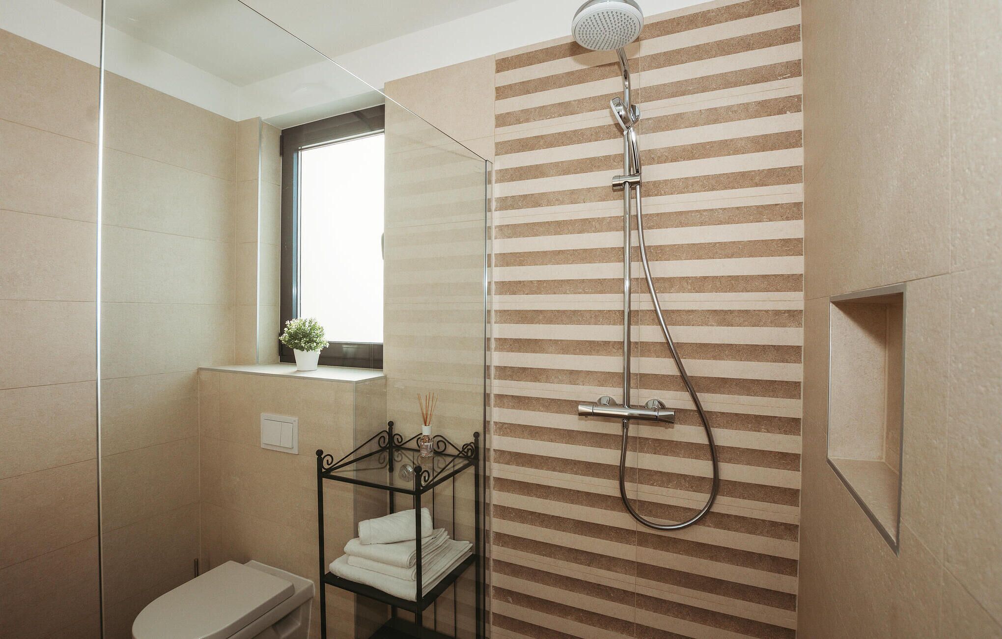 A modern bathroom with a shower, a toilet, a glass shelf with towels, and a window.