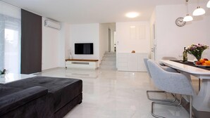 Ein modernes Wohnzimmer mit Flachbildfernseher, einem dunklen Sofa, einem Essbereich mit grauem Stuhl und einer Küche mit weißer Arbeitsplatte.