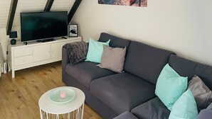 Una sala de estar moderna con un sofá gris, un televisor de pantalla plana, una pequeña mesita blanca con una vela rosa y un mueble blanco.
