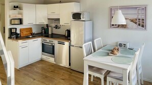 Cocina compacta con gabinetes blancos, microondas, tostadora y cafetera. Hay una zona de comedor con mesa, sillas y radio.