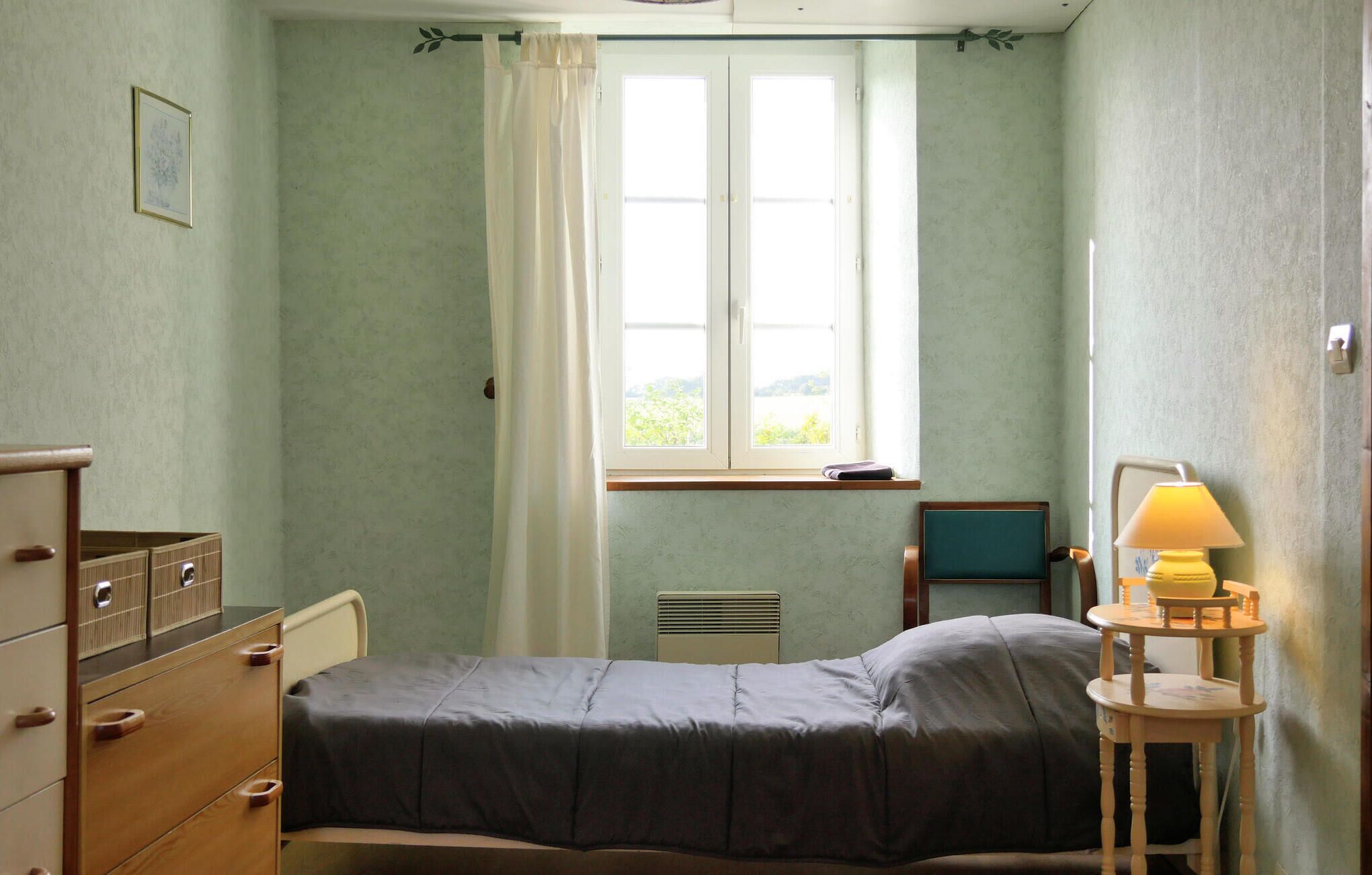 A small, simple bedroom with a single bed, a dresser, a chair, and a bedside table with a lamp.