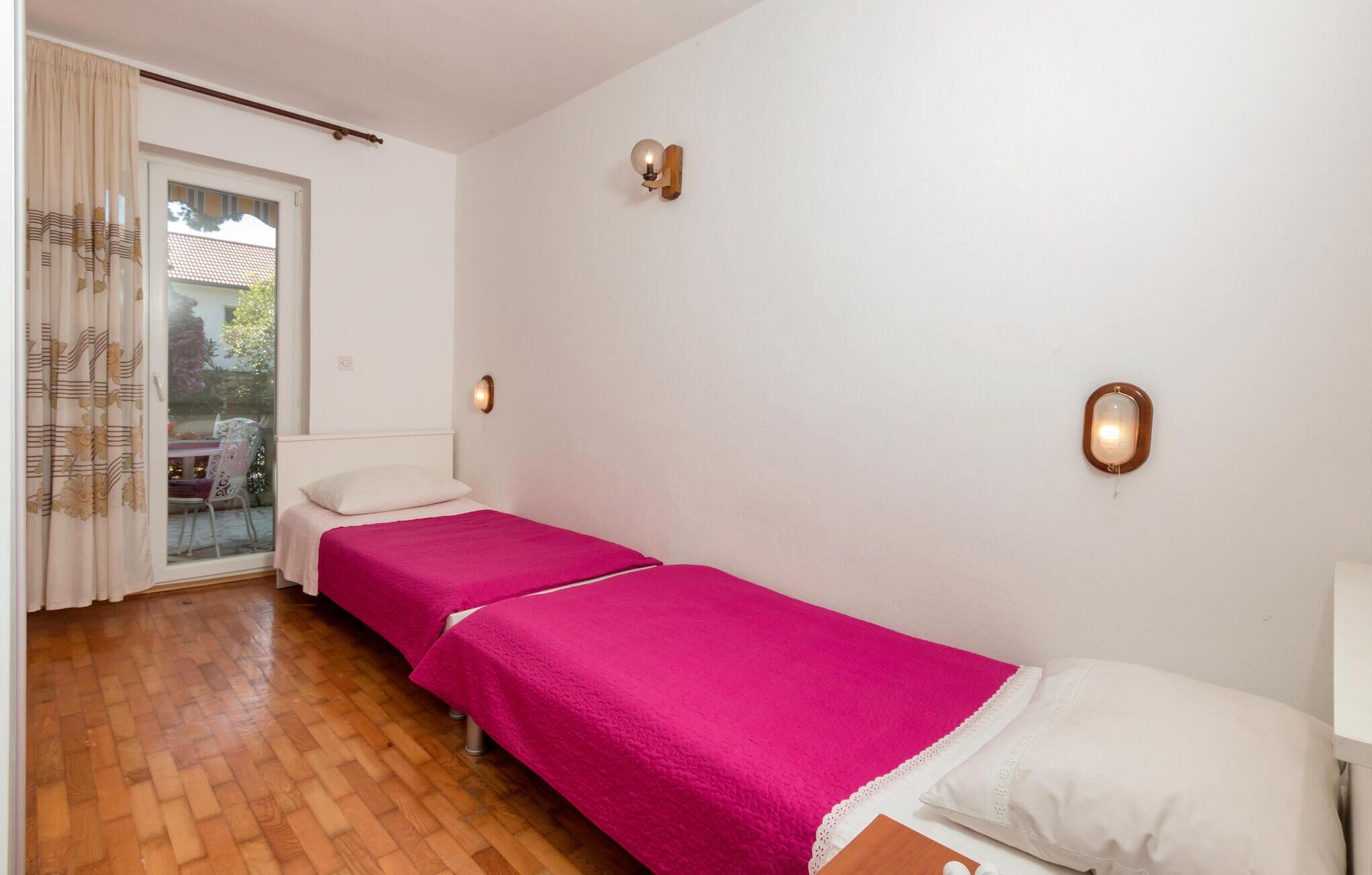 A room with two single beds, a wooden floor, a sliding glass door, and two wall-mounted lights.
