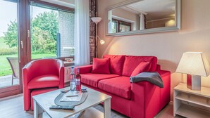 Ein modernes Wohnzimmer mit einem roten Sofa, einem passenden Sessel, einem Couchtisch und einer Lampe. Ein großes Fenster mit Vorhängen, ein Spiegel an der Wand und Blick ins Grüne sind ebenfalls vorhanden.