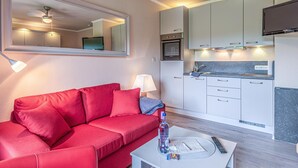 Ein kompakter Wohnbereich mit einem roten Sofa, einem kleinen Couchtisch, einem Fernseher, einer Küchenzeile und einem Spiegel, der den Deckenventilator und die Lampen reflektiert.