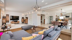 Ein modernes Wohnzimmer mit einem grauen Ecksofa, einem Fernseher, Holzschränken und einem Essbereich mit Tisch und Stühlen.