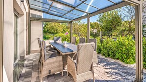Eine überdachte Terrasse mit Tisch und Stühlen, umgeben von Grünpflanzen und blauem Himmel.