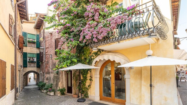 A narrow cobblestone alley with a building featuring a balcony with purple flowers, a wooden door, and a white awning.