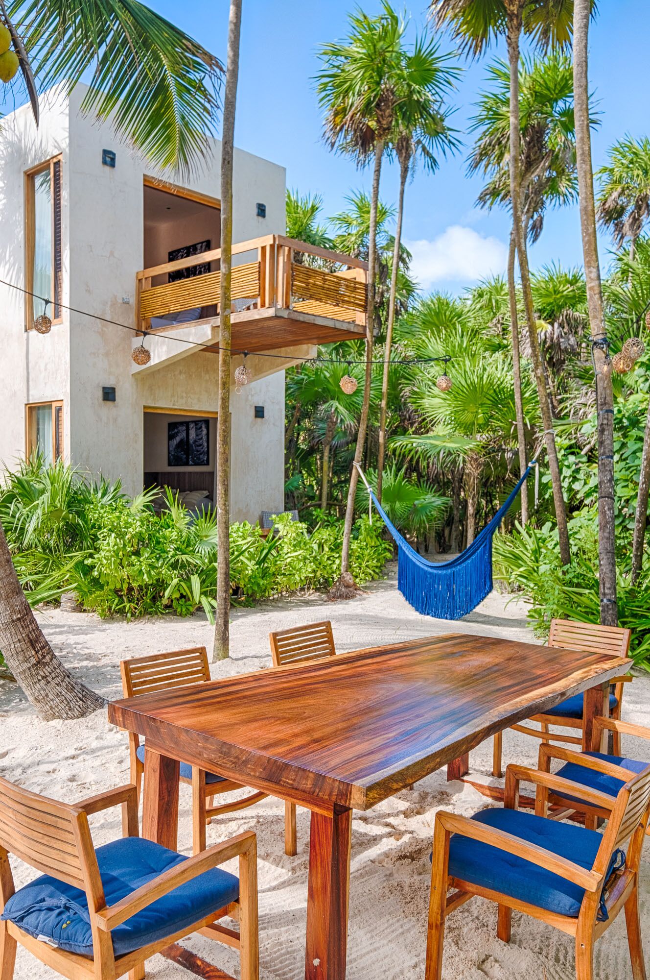 A wooden dining table with blue cushions, surrounded by chairs, set on a sandy area with a hammock and lush greenery in the background.