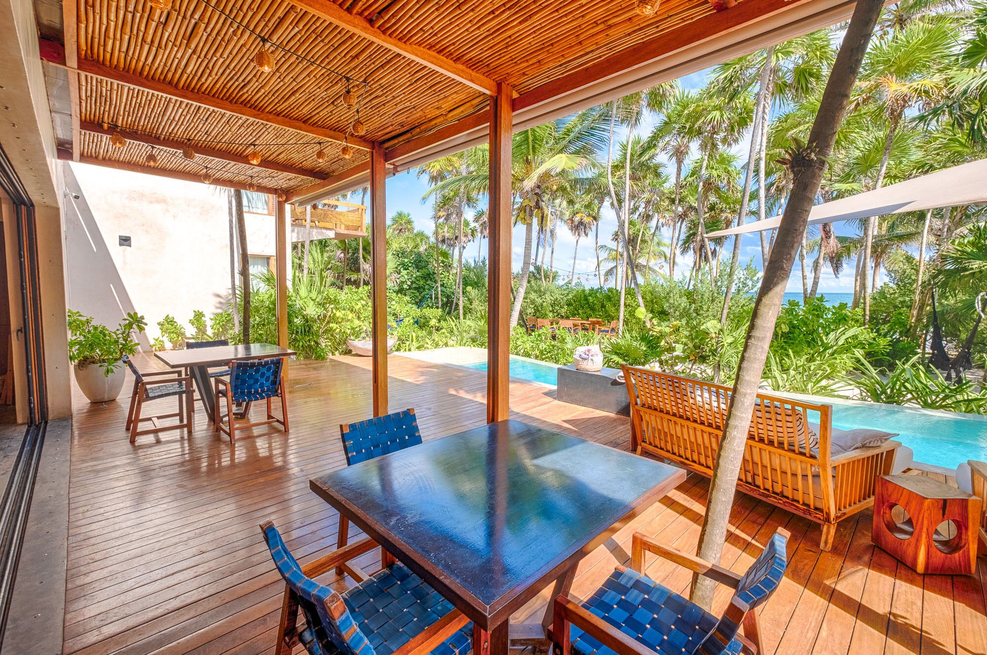 A covered outdoor area with a pool, wooden furniture, and tropical plants.