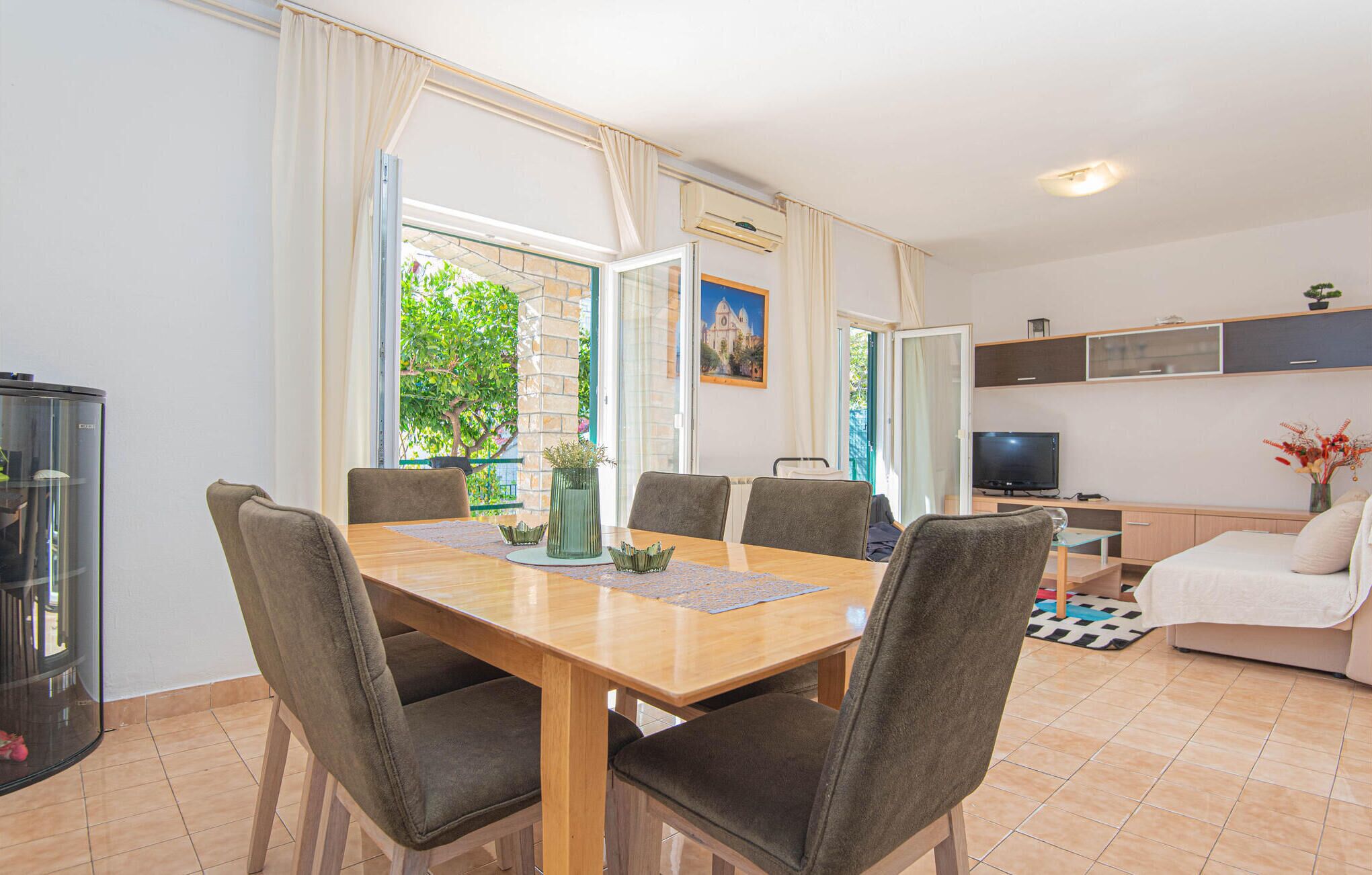 Uma área de refeições com mesa de madeira e seis cadeiras, televisão, sofá e cozinha com armários e micro-ondas.