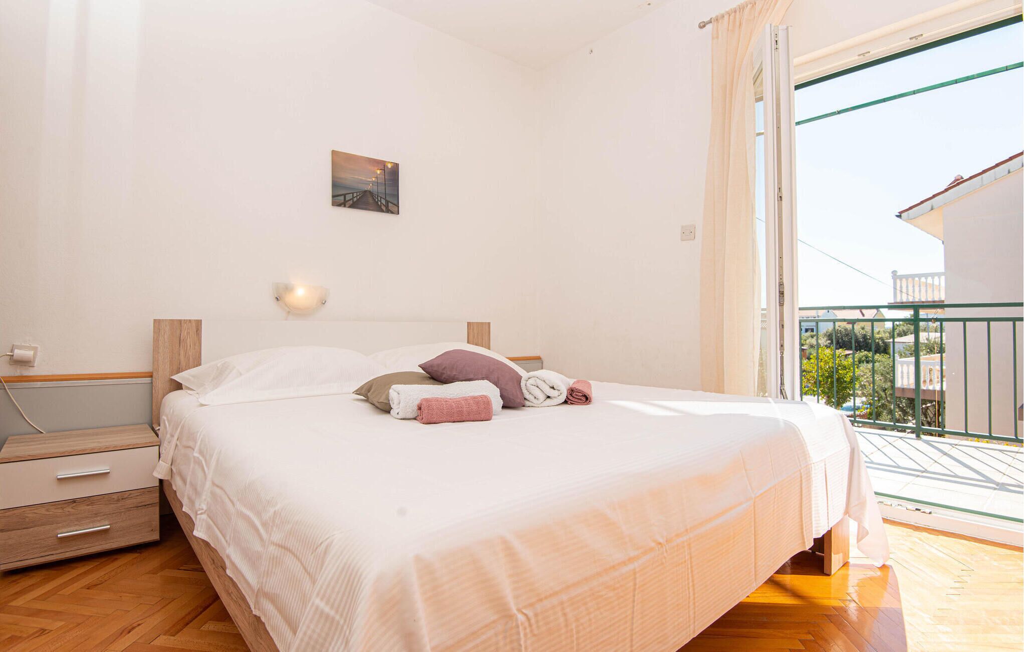 A bedroom with a wooden headboard, a single bed with white bedding, a nightstand with a lamp, and a view of the outside balcony.
