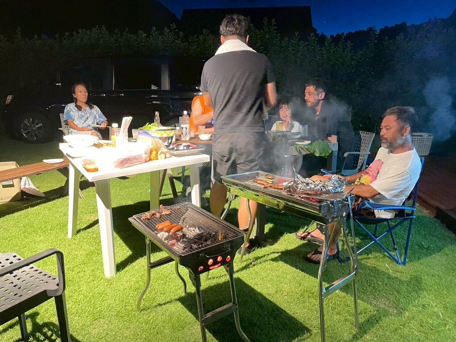 An outdoor barbecue with people grilling and sitting around a table with food and drinks.