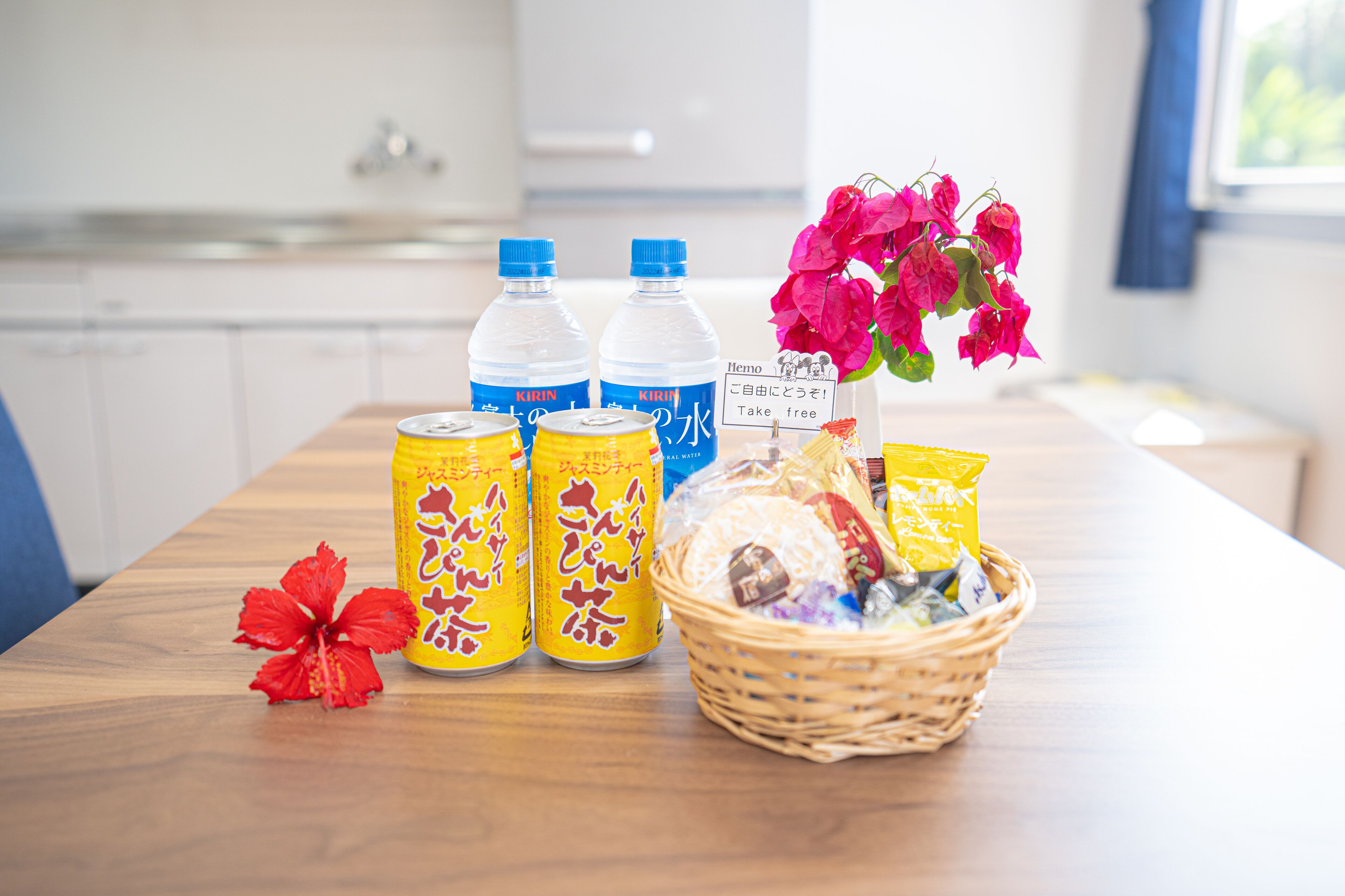 A wooden table with three cans of Kirin beer, a basket of snacks, and a red hibiscus flower.