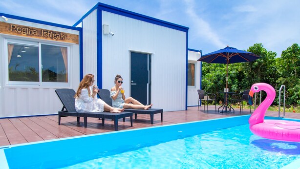 Una piscina con sillones de descanso, un flotador con forma de flamenco rosa y un edificio con un letrero que dice "Regina Ishigaki".