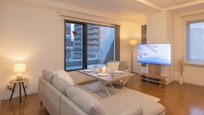 Ett modernt vardagsrum med en soffa, ett matbord för två personer, en tv och ett stort fönster med utsikt över staden.