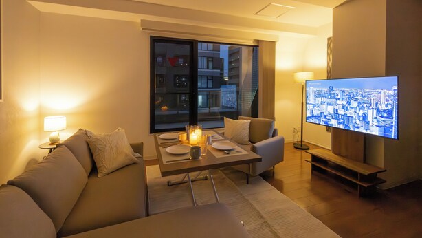 Ett modernt vardagsrum med en soffa, ett matbord dekorerat med ljus, en tv-skärm som visar en stadsbild och ett stort fönster med utsikt över byggnader.