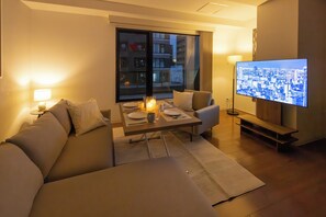 A modern living room with a sofa, a dining table set with candles, a TV displaying a cityscape, and a large window with a view of buildings. - Atrium - Self Check-In Only (Osaka)