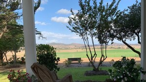 Un porche cubierto con una mecedora, plantas en macetas y un banco con vistas a un campo verde y colinas al fondo.