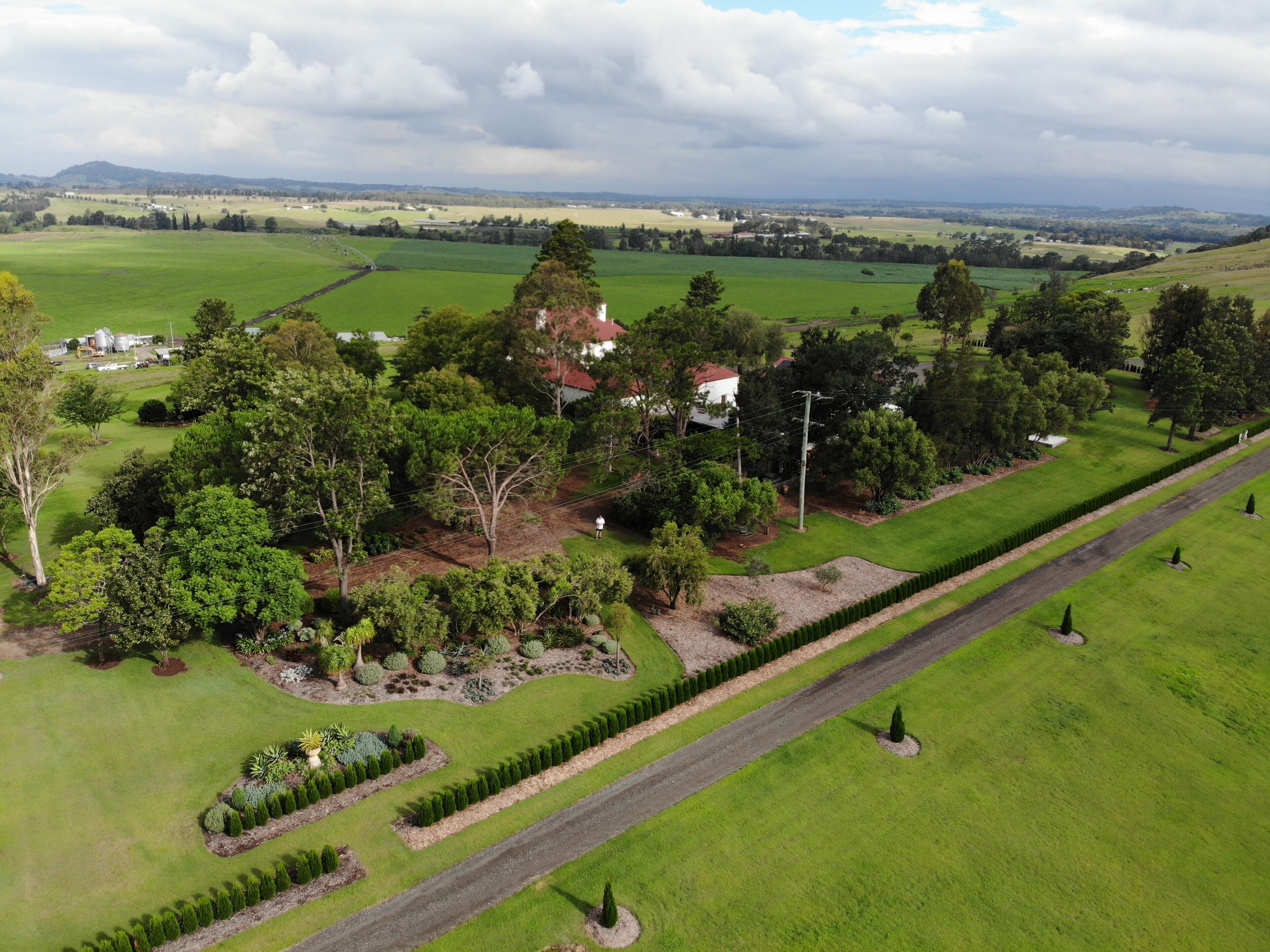 An aerial view of a property with a well-maintained lawn, a variety of trees, and a road running through the center.
