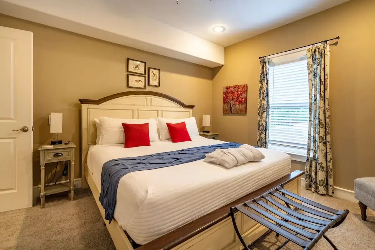 A hotel room with a large bed, a bench, a nightstand, a window with curtains, and artwork on the wall.