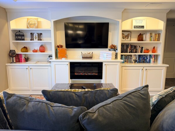 Ein Wohnzimmer mit Flachbildfernseher über einem Kamin, weißen Schränken und einem Bücherregal mit Büchern und Dekoration.