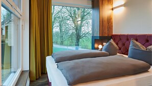 Une chambre d’hôtel dotée d’une grande fenêtre, d’un lit avec une literie grise, d’une tête de lit rouge rembourrée et d’un luminaire fixé au mur.