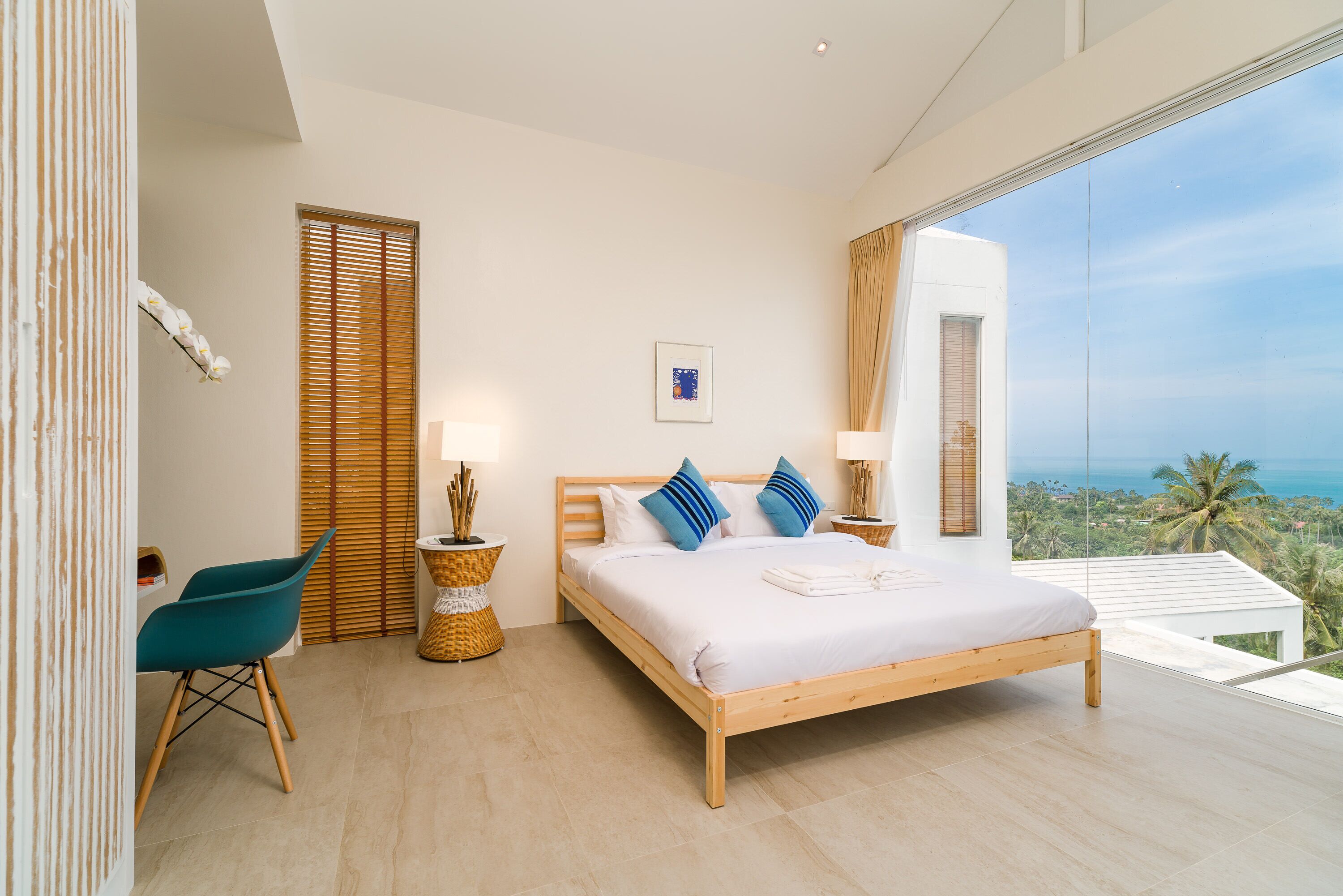 Een moderne slaapkamer met een houten bed, uitzicht op de oceaan, een rieten stoel en een tafeltje met een lamp.