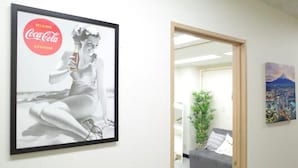A framed Coca-Cola advertisement on a wall, a gray sofa with a plant, and a colorful painting in a room.