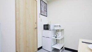 A small, clean room with a wooden door, a white refrigerator, a microwave, and a white shelving unit with a kettle. - TORA HOTEL Taito B (Tokyo)