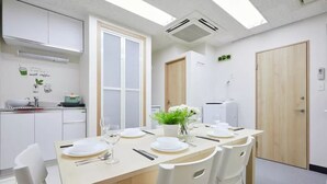 A modern dining area with a wooden table set for four, white chairs, a small kitchen area with a sink and cabinets, and a door with a green plant decoration. - TORA HOTEL Taito B (Tokyo)