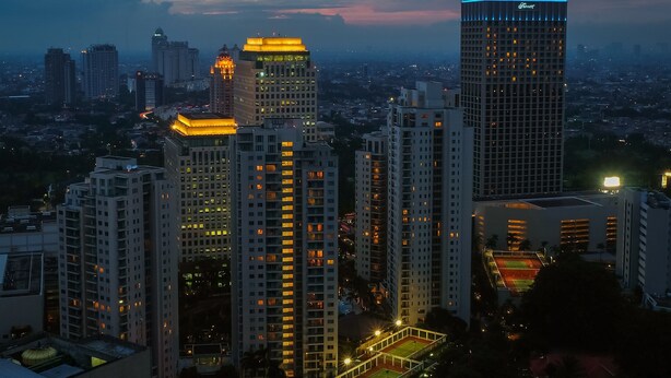A cityscape at dusk with high-rise buildings, one with a distinctive blue-lit top. - Fantastic View 2BR Apartment at FX Residence Sudirman (Jakarta)