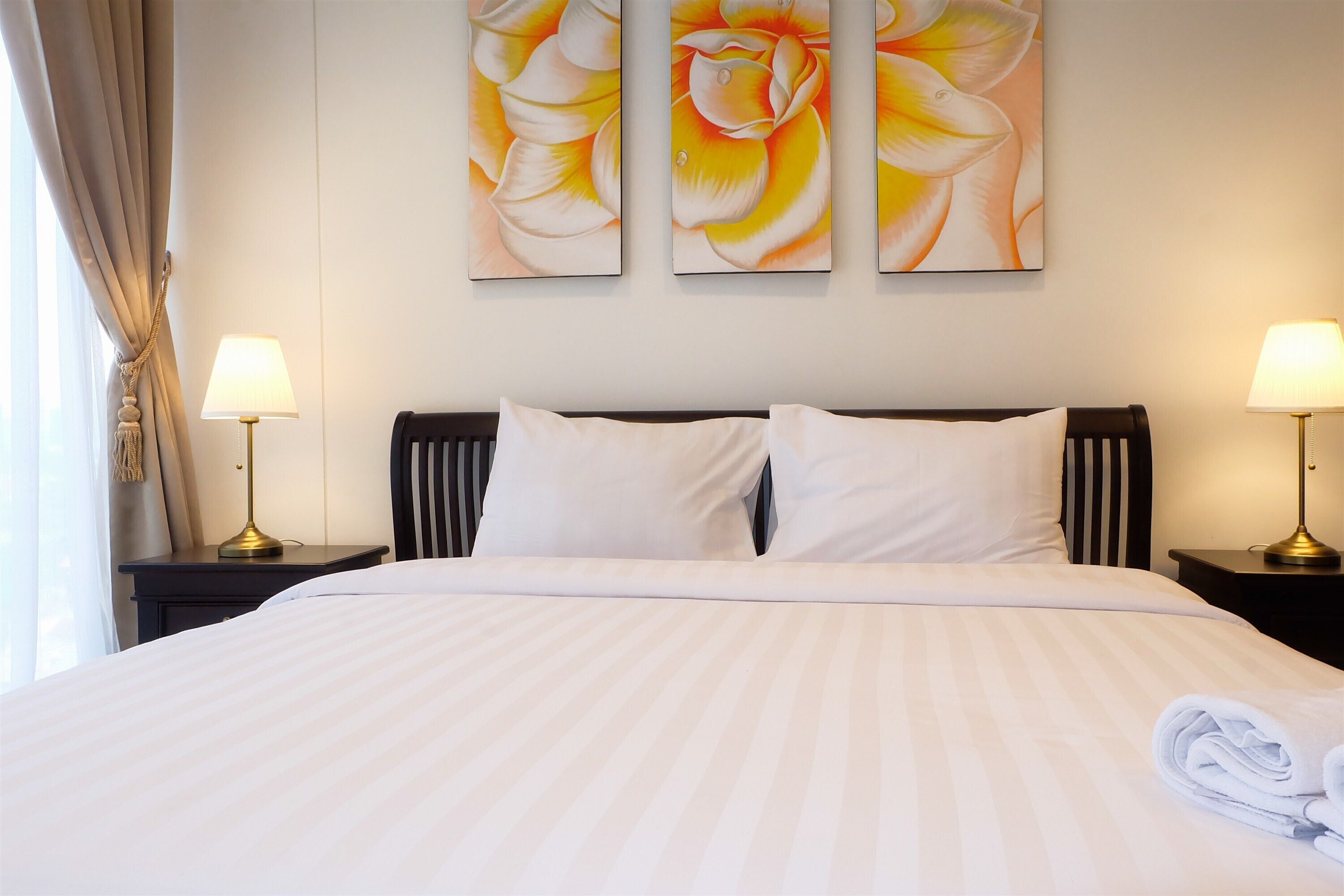 Una cama bien hecha con sábanas blancas, dos lámparas de noche y una gran obra de arte floral sobre la cama.