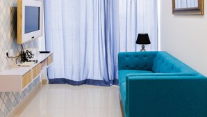 A modern bedroom with a blue sofa, a TV on a wooden stand, a lamp, and a large window with curtains. - Nice and Prime Location 1BR Casa De Parco Apartment (Pagedangan)