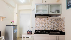 Cocina compacta con refrigerador, estufa y lavamanos. Hay una pequeña mesa con un taburete rojo y una planta en maceta.