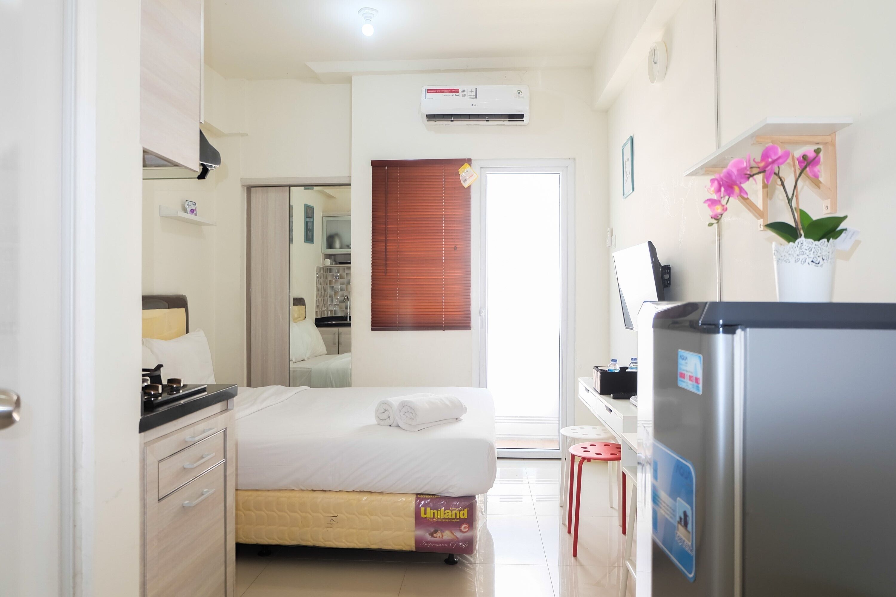A compact hotel room with a bed, a kitchenette, a refrigerator, and a window with blinds.