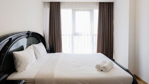 A hotel room with a large bed, a black headboard, white bedding, and two windows with brown curtains. - Nice 1BR Apartment at M-Town Signature (Gading Serpong)