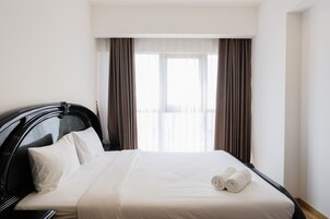 Una habitación de hotel con una cama grande, un cabecero negro, ropa de cama blanca y dos ventanas con cortinas marrones.