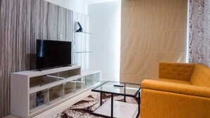A modern living room with a flat-screen TV on a white entertainment unit, a glass coffee table, and a mustard-colored sofa. - Premium 1BR Apartement At Grand Sungkono Lagoon (Surabaya)