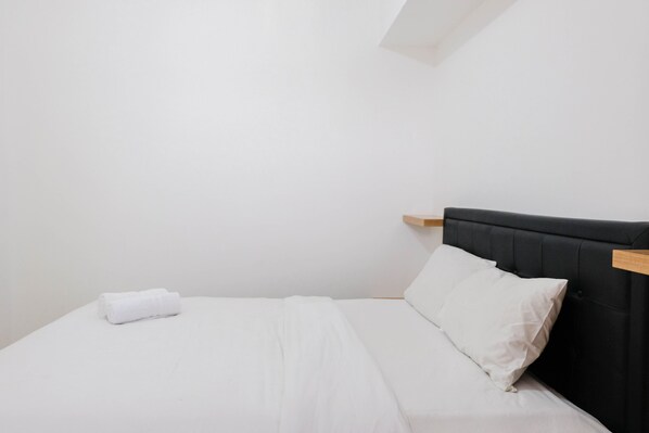 Un lit bien fait, avec du linge de lit blanc et une seule serviette enroulée, une tête de lit noire recouverte de tissu et une étagère en bois.