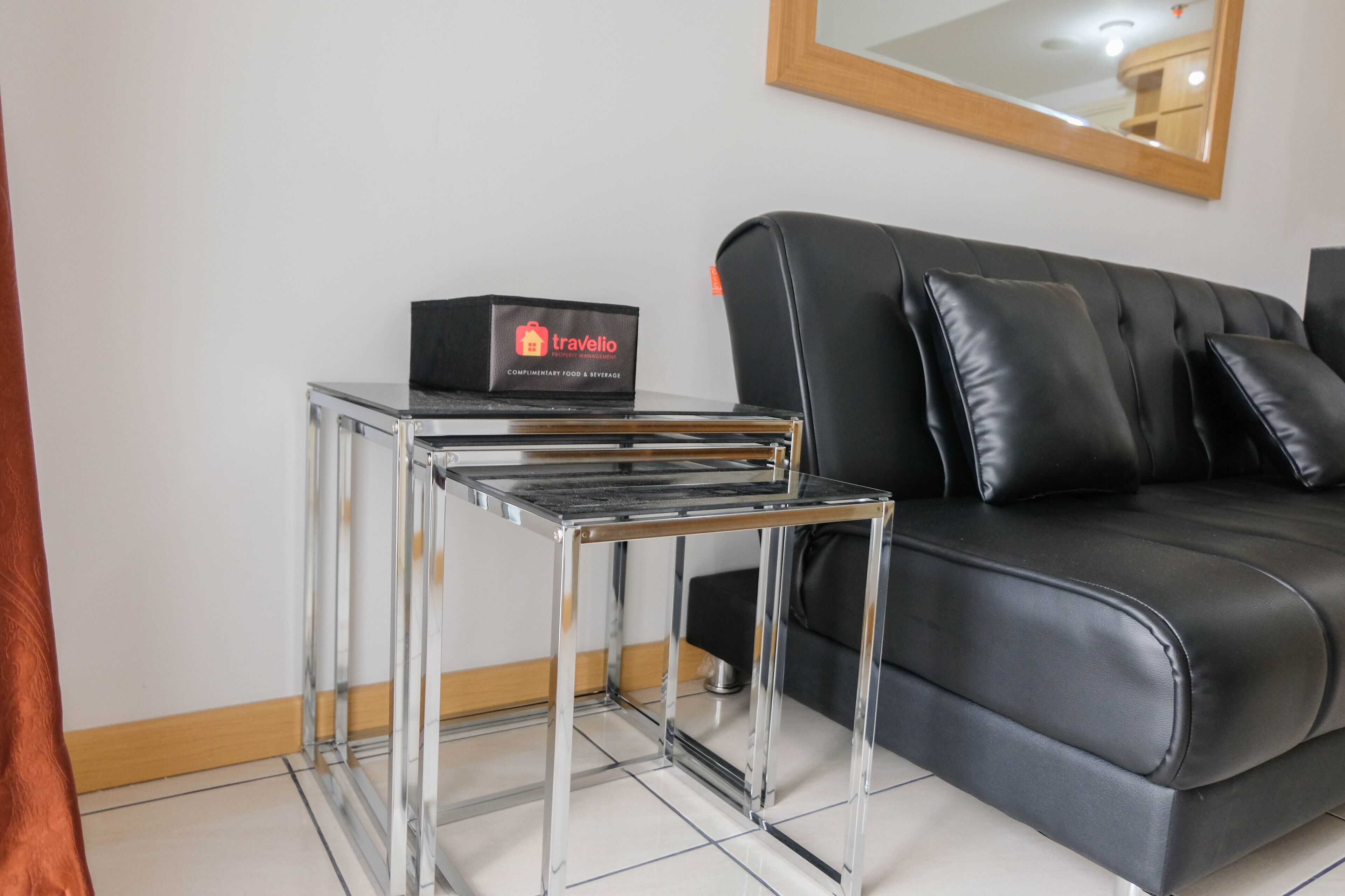 A modern living room with a black leather sofa, a chrome side table, and a black box labeled 'Travelio'.