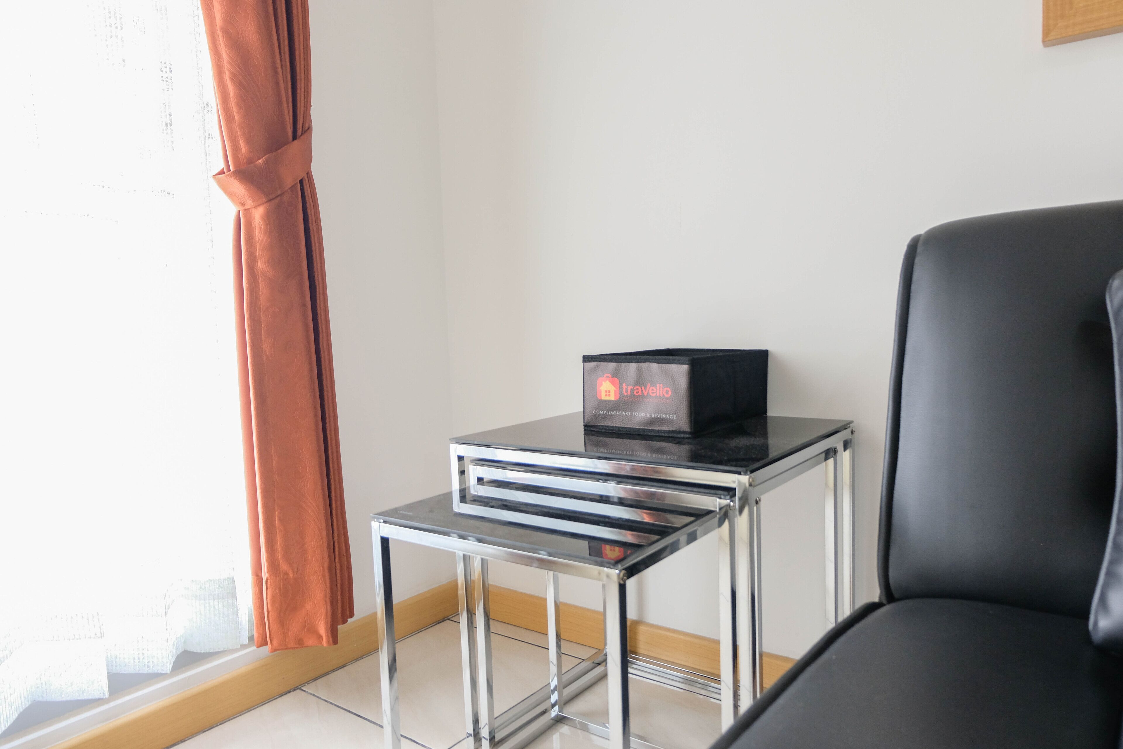 A modern room with a black leather chair, a small glass table with a black box labeled "travelio," and a window with orange curtains.