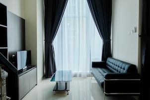 A modern living room with a flat-screen TV on a cabinet, a glass coffee table, a black leather sofa, and sheer curtains allowing natural light.