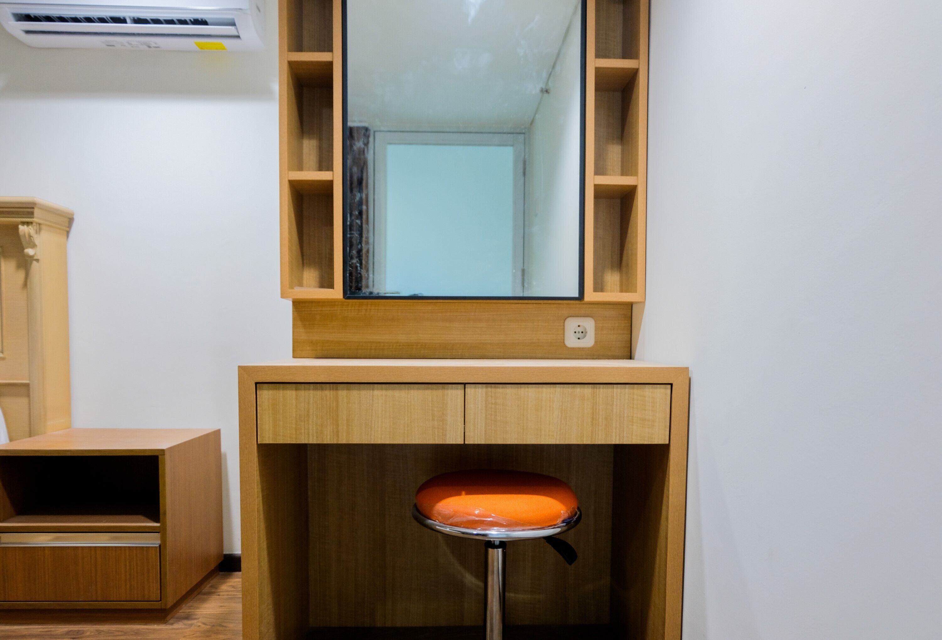 A room with a wooden desk, a stool, a mirror, and an air conditioning unit.
