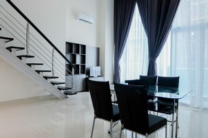 Una habitación de hotel moderna con una mesa de comedor de cristal, sillas negras, una escalera y un amplio ventanal con cortinas.