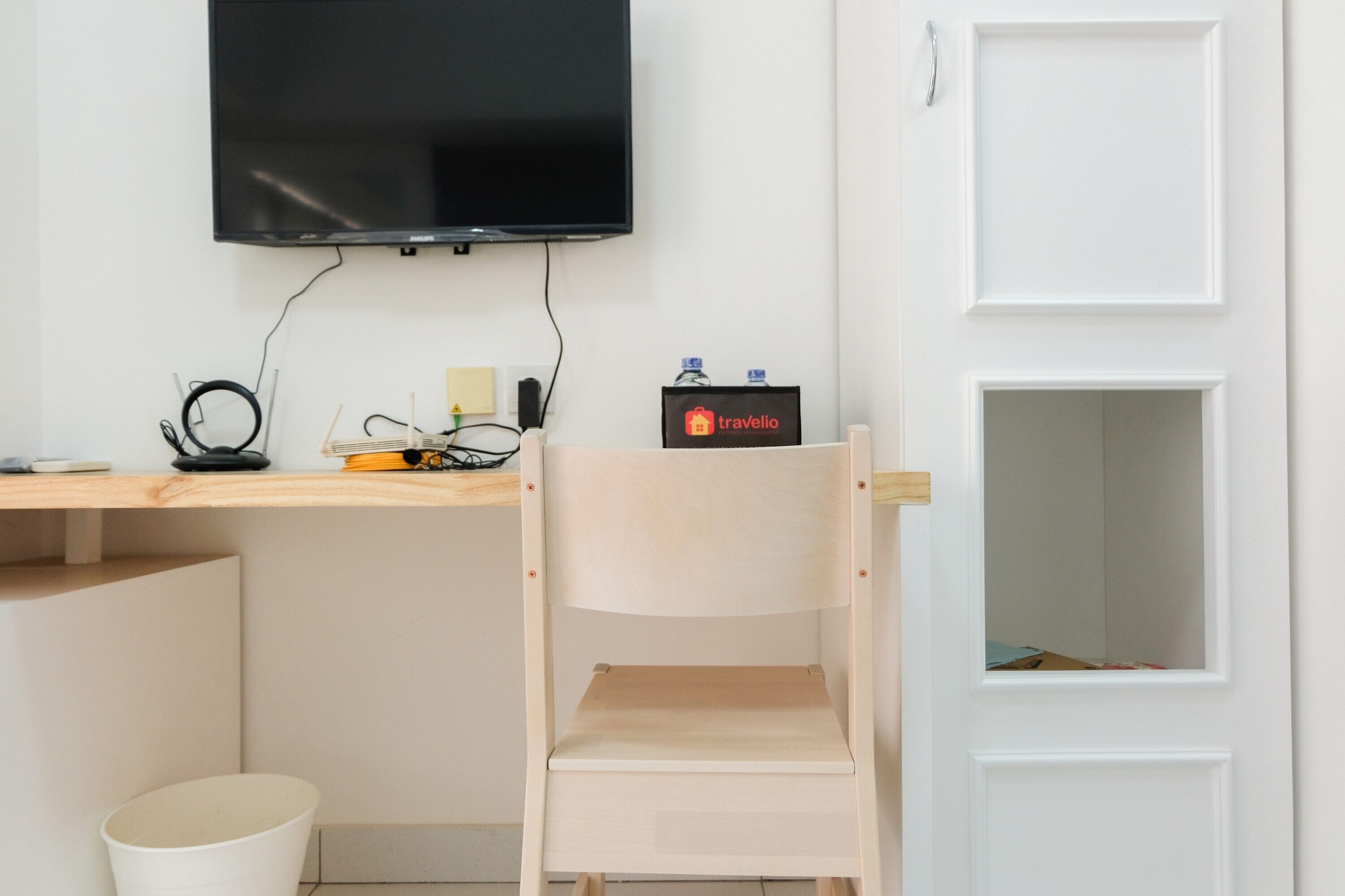A room with a wooden desk, a chair, a TV mounted on the wall, a door, and a small shelf with a water bottle and a bag.