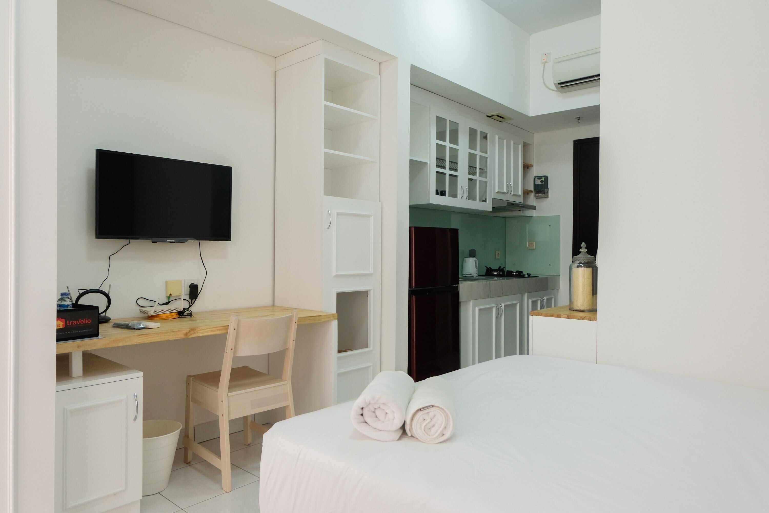 A hotel room with a bed, a desk with a chair, a television, a kitchen area, and a minibar.
