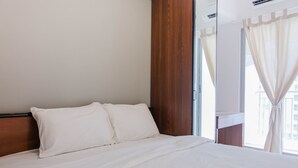 A hotel room with a large bed, wooden headboard, and a view of the city through sheer curtains. - Comfortable 2BR Apartment Serpong M-Town Residence (Gading Serpong)