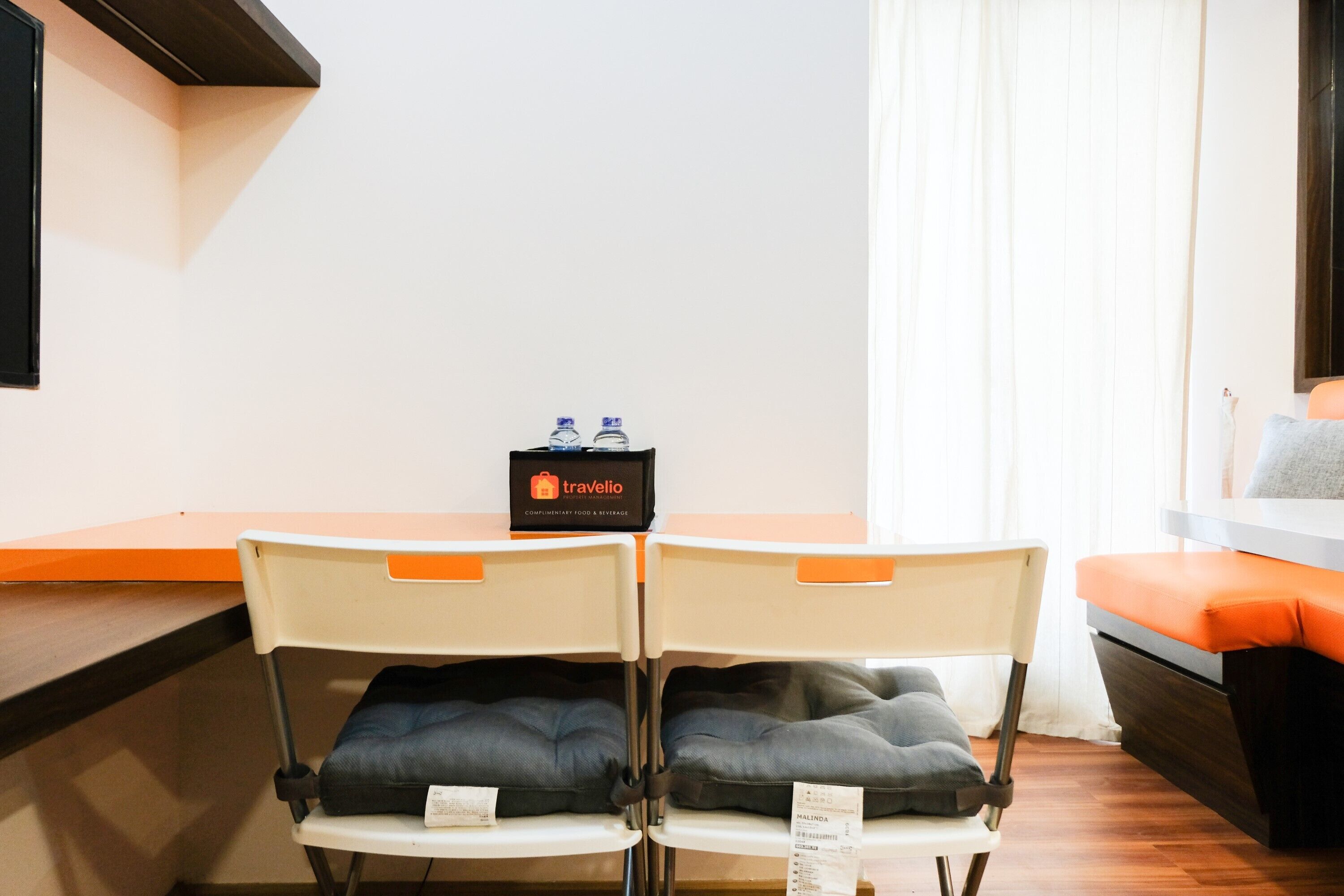 A room with two chairs, a small table, a bed with orange bedding, a window with curtains, and a travel bag on the table.