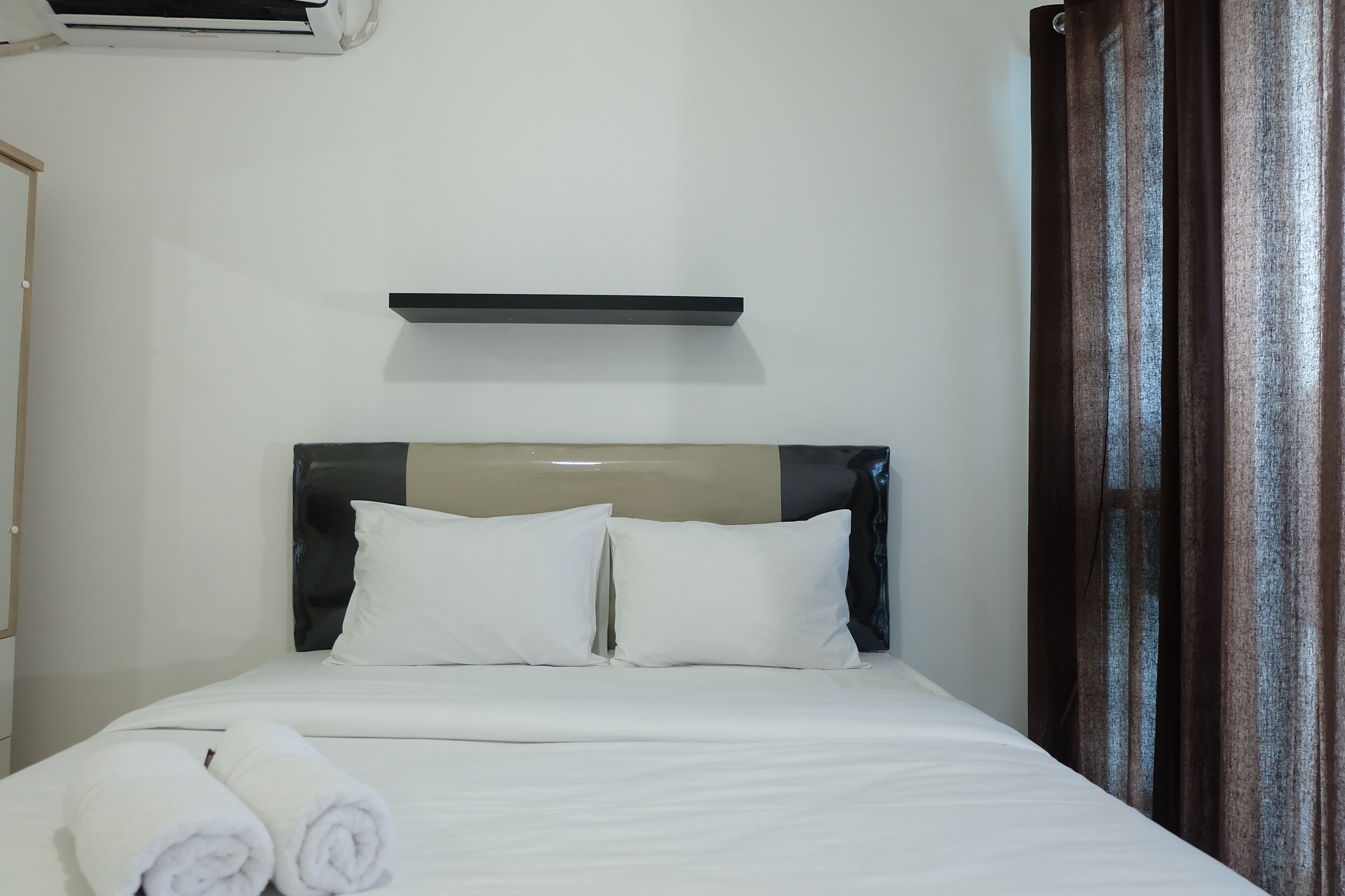 A hotel room with a bed, white pillows, a headboard, a small shelf above the bed, and a curtain on the right side.